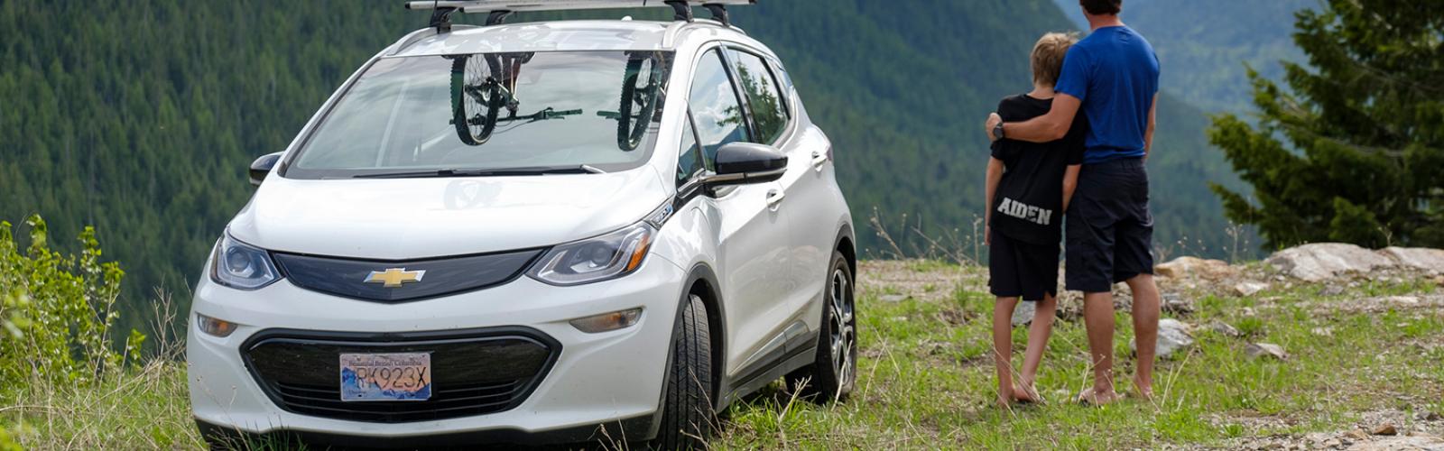 An electric vehicle, bearing two mountain bikes on the roof rack, parked atop a lookout beside a young couple embracing and taking in a stunning landscape of mountains of trees