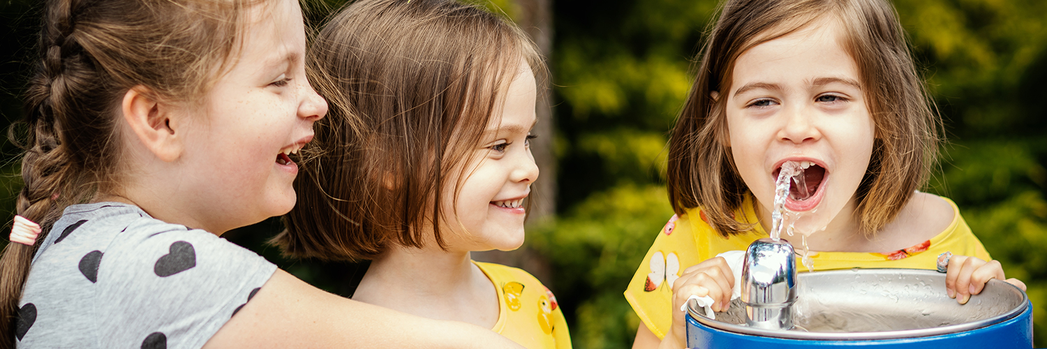 girl-in-a-yellow-dress-with-sisters-drinks-water-from-a-fountain