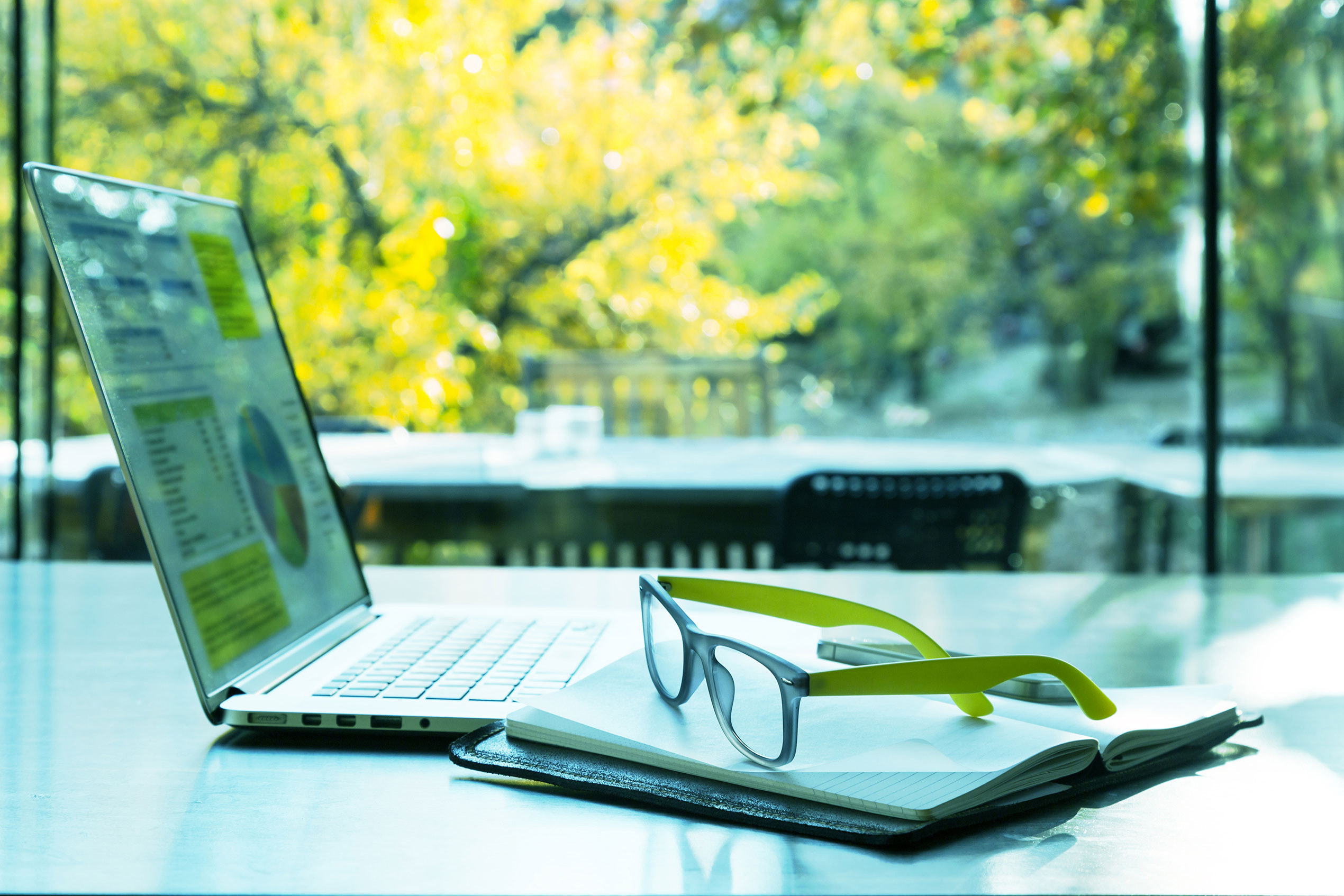 Laptop sits on glass table beside a pair of reading glasses. 