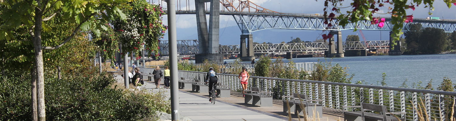 A boardwalk park in New Westminster featuring trees, greenery, park benches, and a bridge in the background