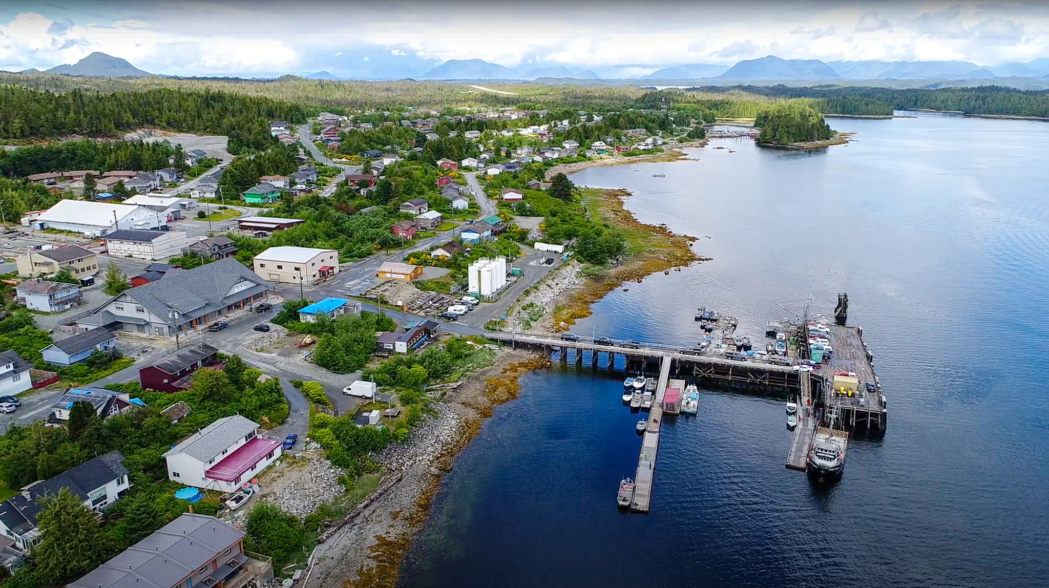 An aerial view of the town of Bella Bella, showing buildings and a dock into the ocean as well as forest and mountains in the background