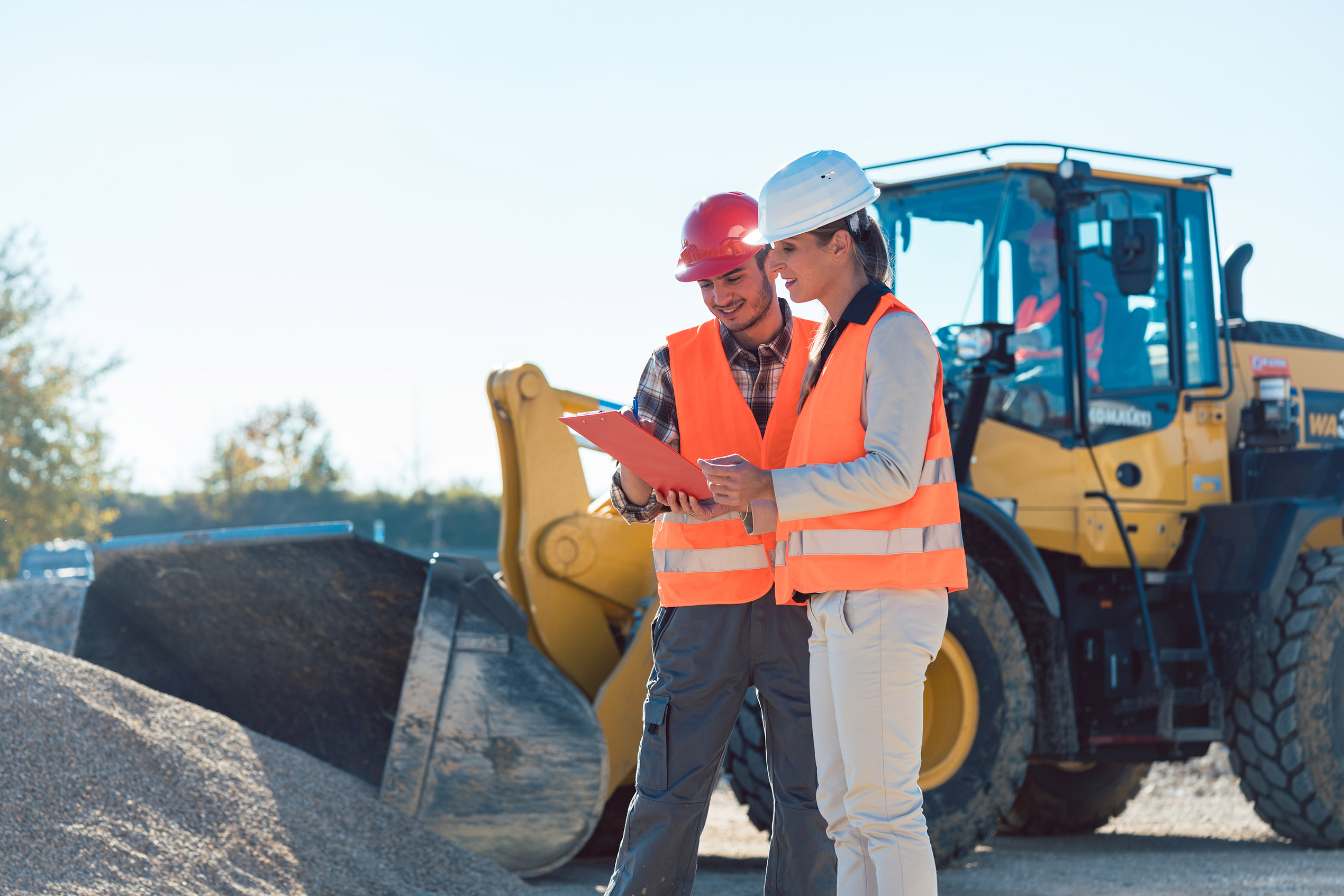 Man and woman in construction gear discussing blueprints while standing on a building site with construction equipment in the background