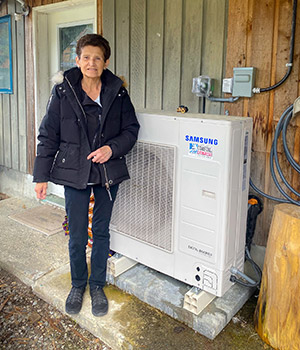 A person standing outdoors next to a house and the outdoor unit of its heat pump system