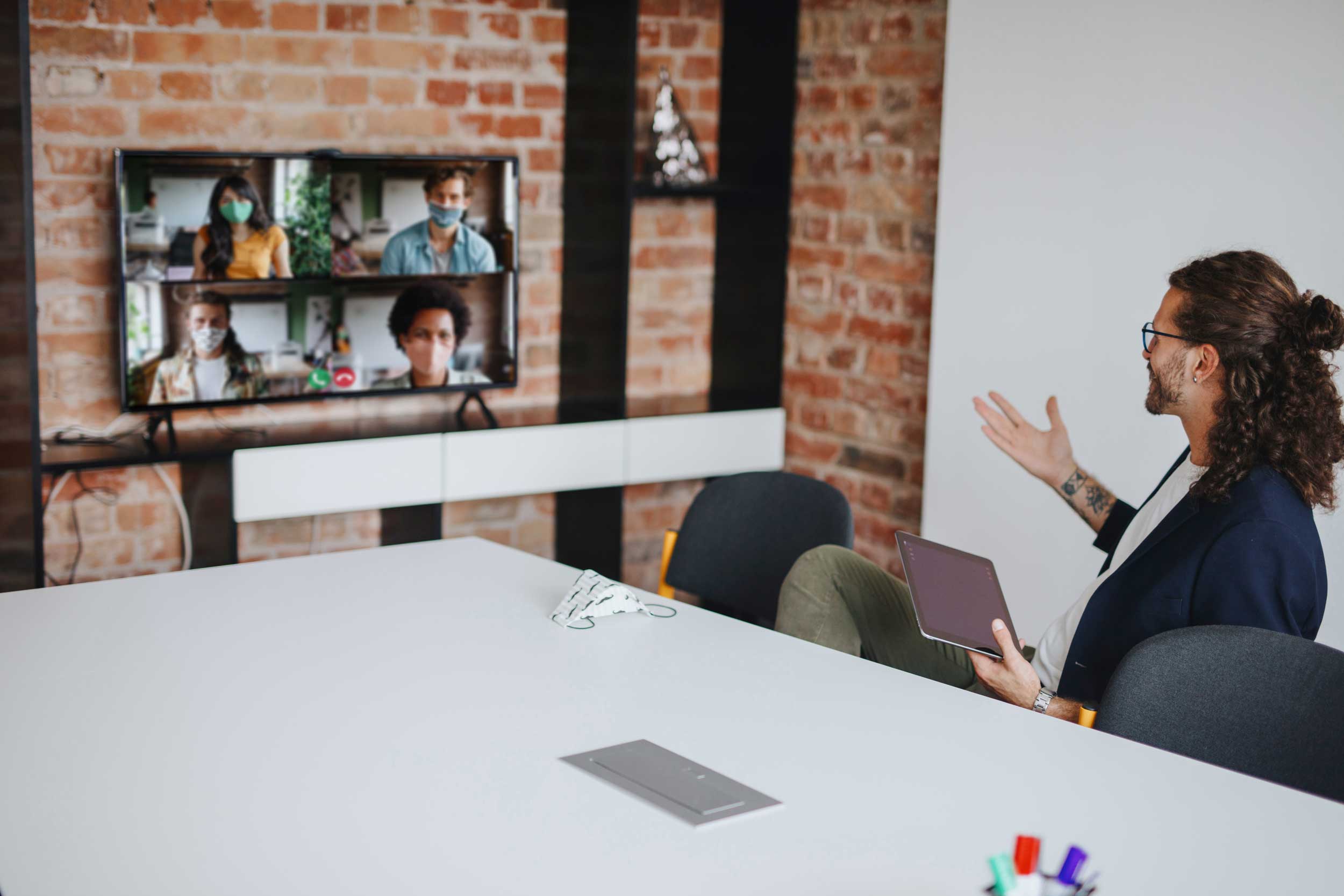 Businessman in an exposed brick office uses a large monitor on the wall to have a video call with several colleagues.