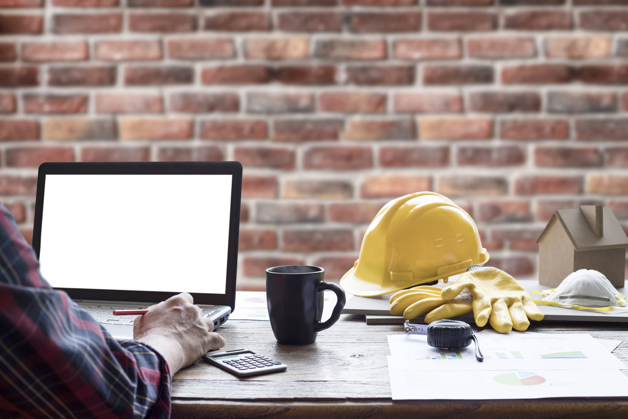 Man uses a laptop sitting at a desk, yellow hardhat set aside. 