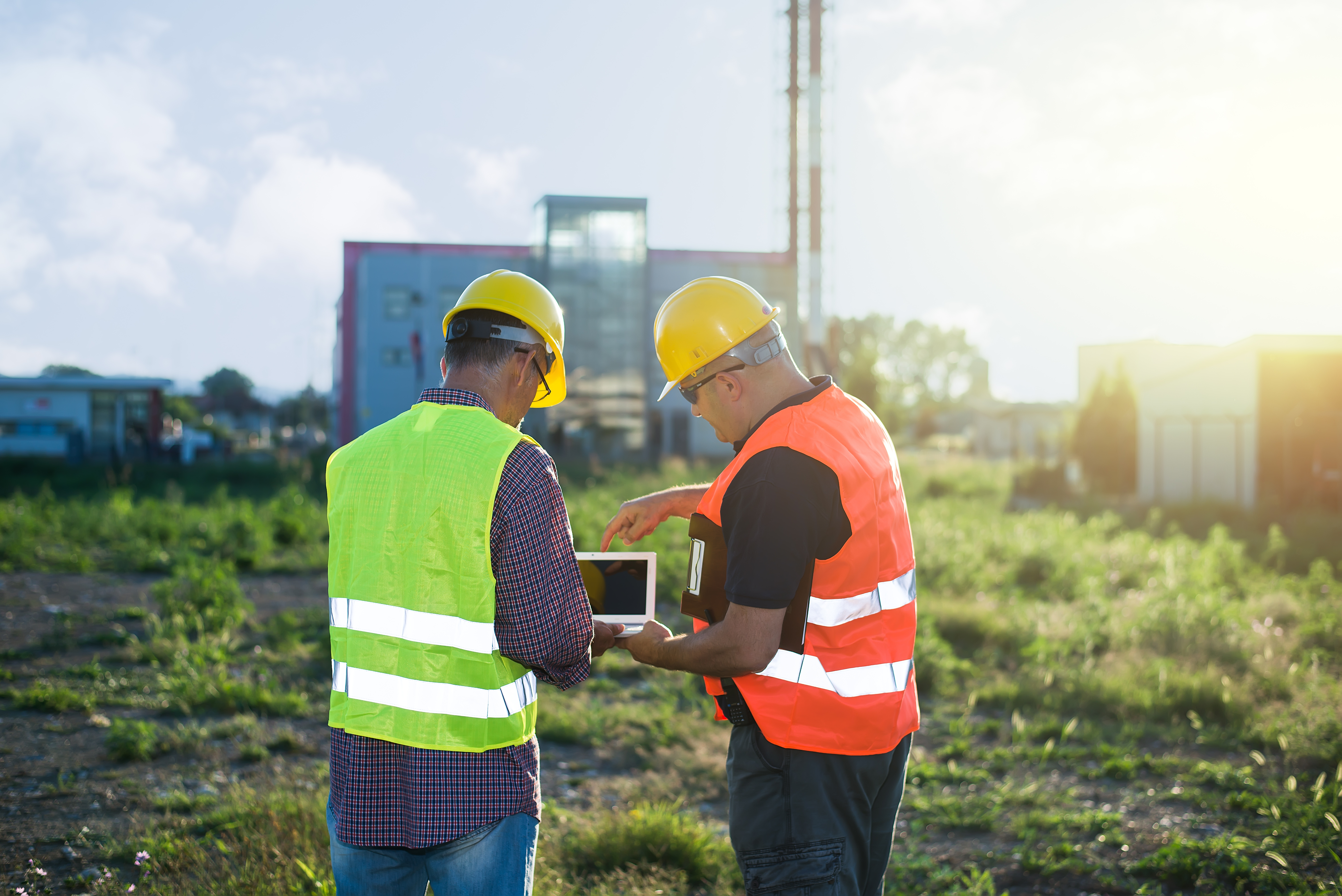 Two engineers in high-visibility safety vests and hard hats face away from the camera as they assess a facility.