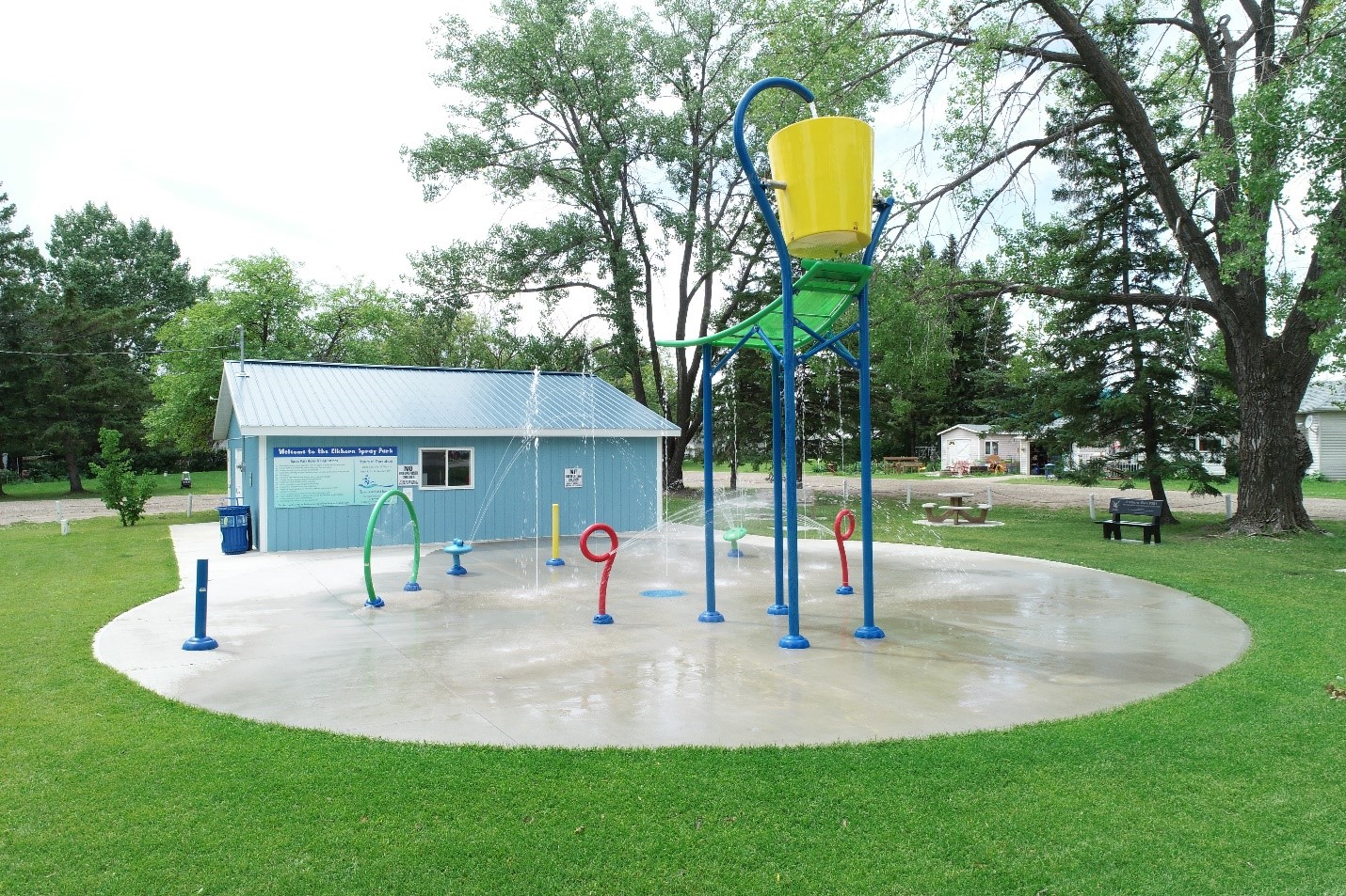 A colourful water spray park with a small blue outbuilding, surrounded by grass and trees.