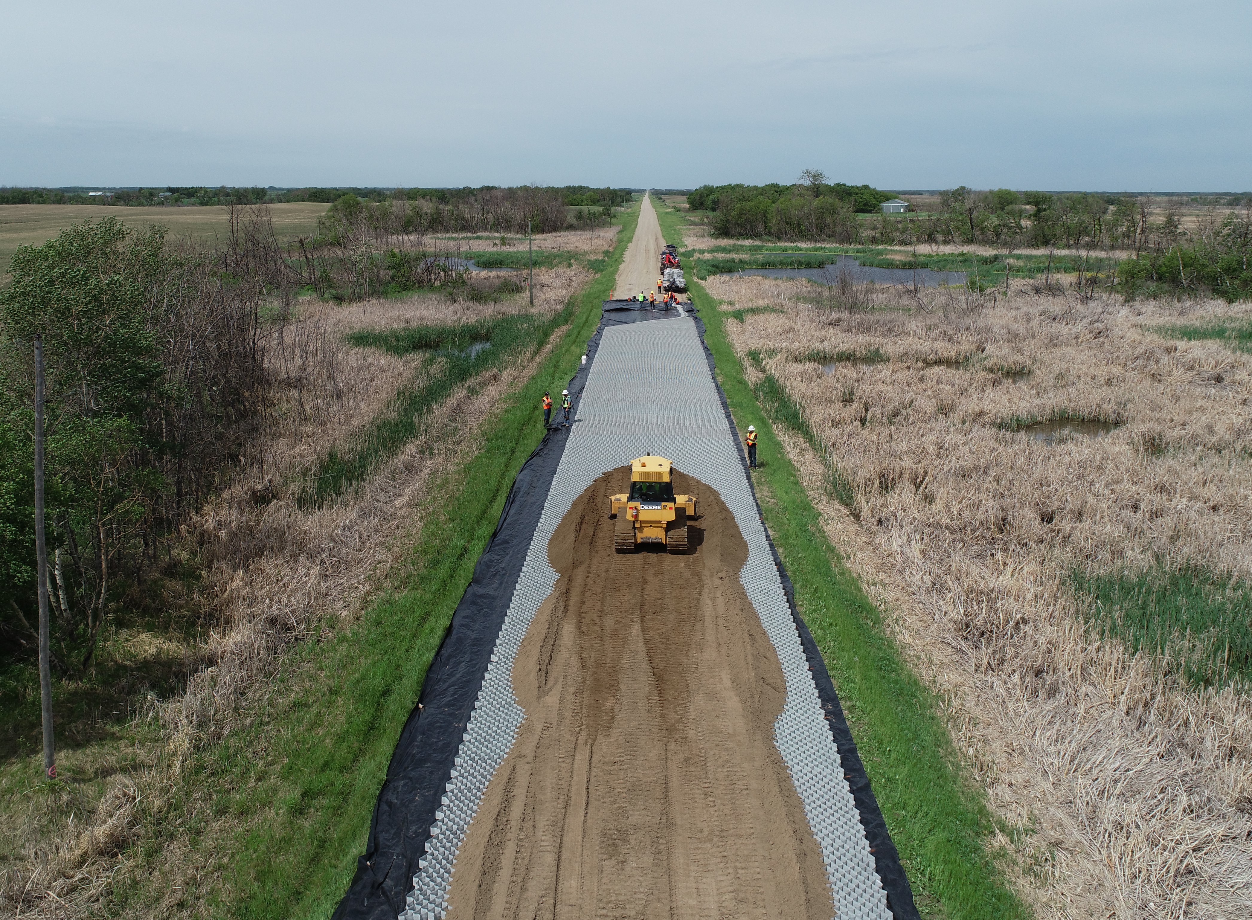 A road crew uses construction equipment to work on a wide gravel road, with fields on both sides
