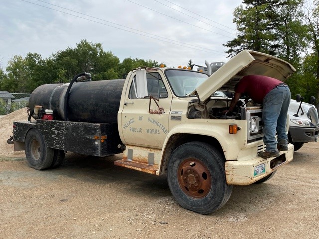 A maintenance worker works under the hood of an old public works truck in a parking lot surrounded by trees.