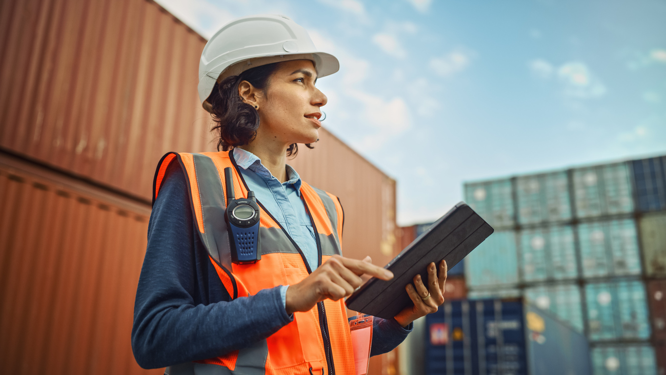 Woman in white hard hat inspects a construction site while holding a tablet.