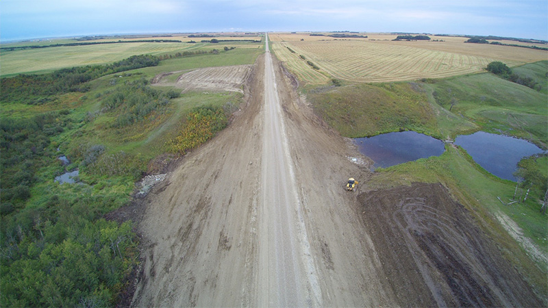 Aerial view of a newly constructed gravel road between green and gold fields with a construction loader near a small slough.