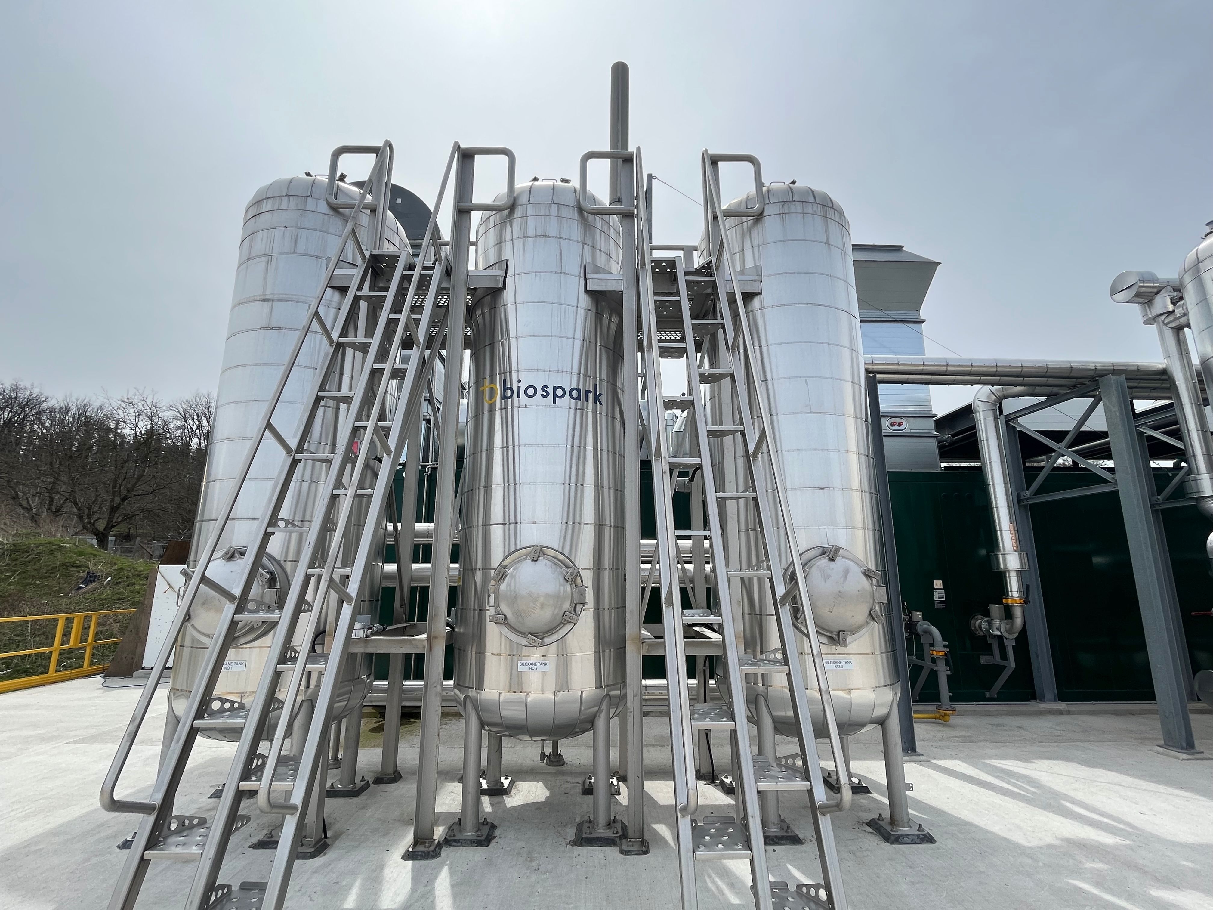 Outdoor view of three tall metal tanks mounted to concrete, with ladders for access.