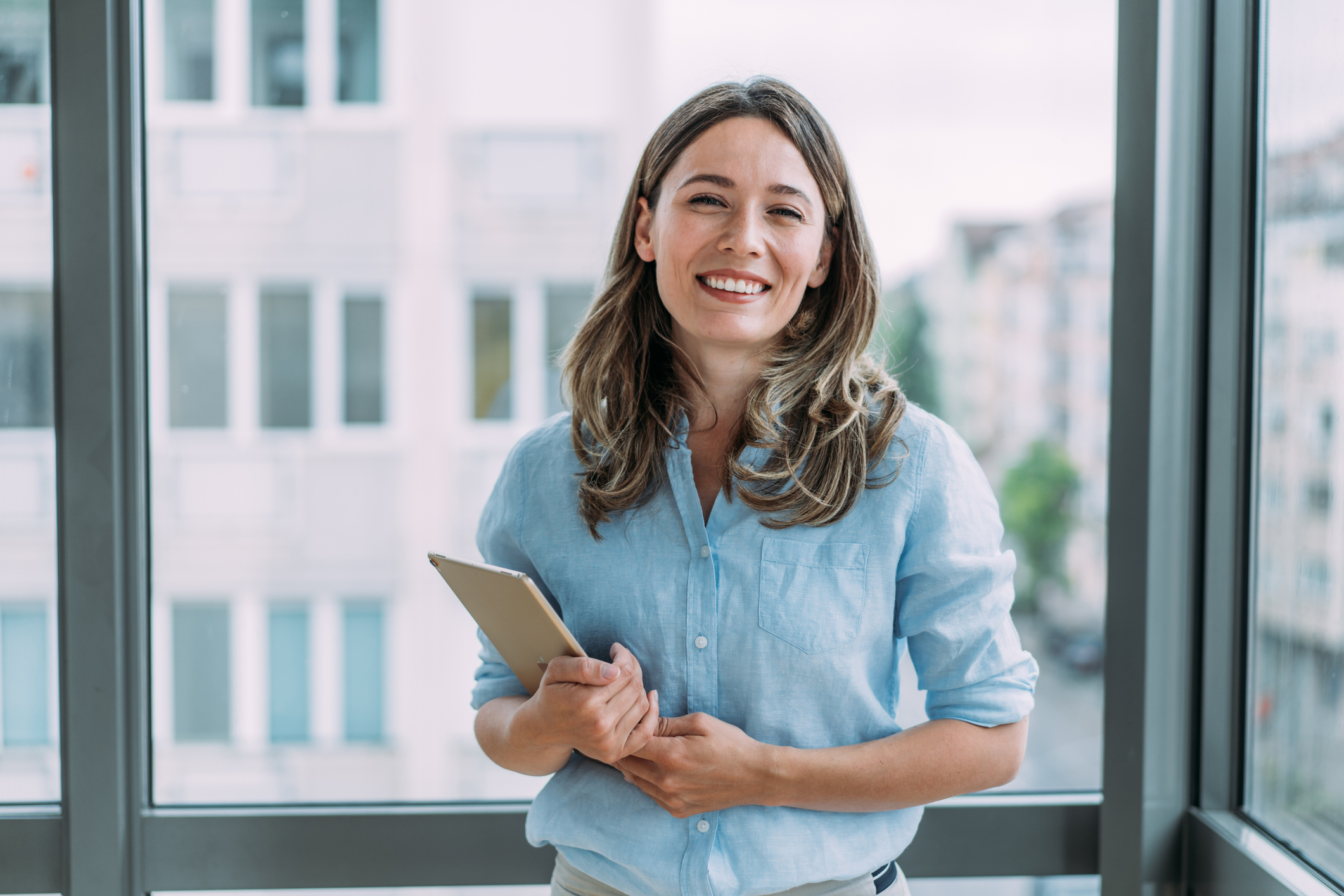 Smiling, confident woman in a bright and sunny office holds paperwork while standing in front of a large window. 