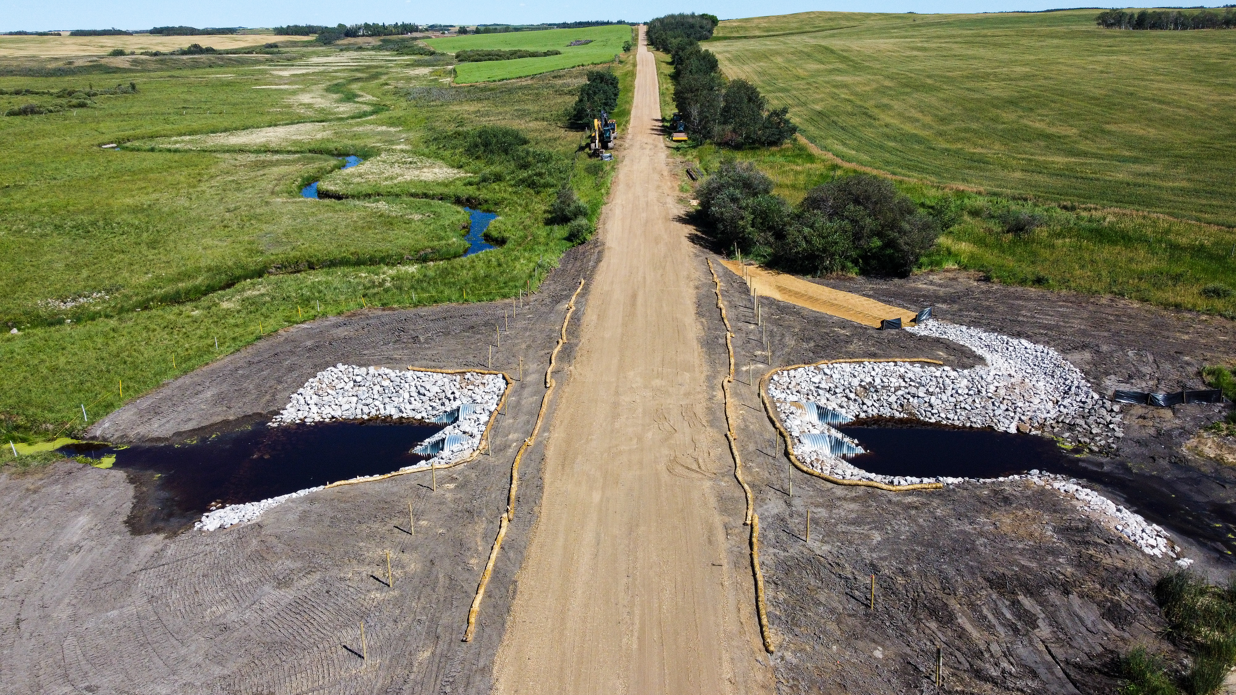 A gravel road across a newly replaced bridge, with soil on both sides, fields and a winding tributary.