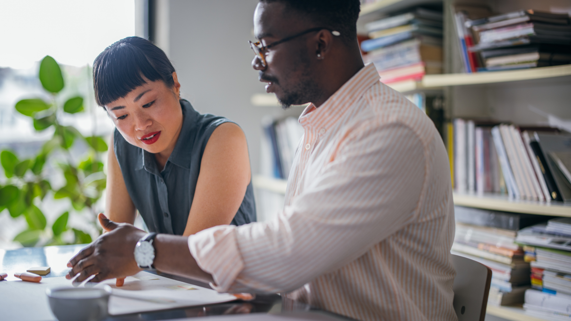 Two colleagues in a bright, sunny and cozy office have a discussion while looking over paperwork. 