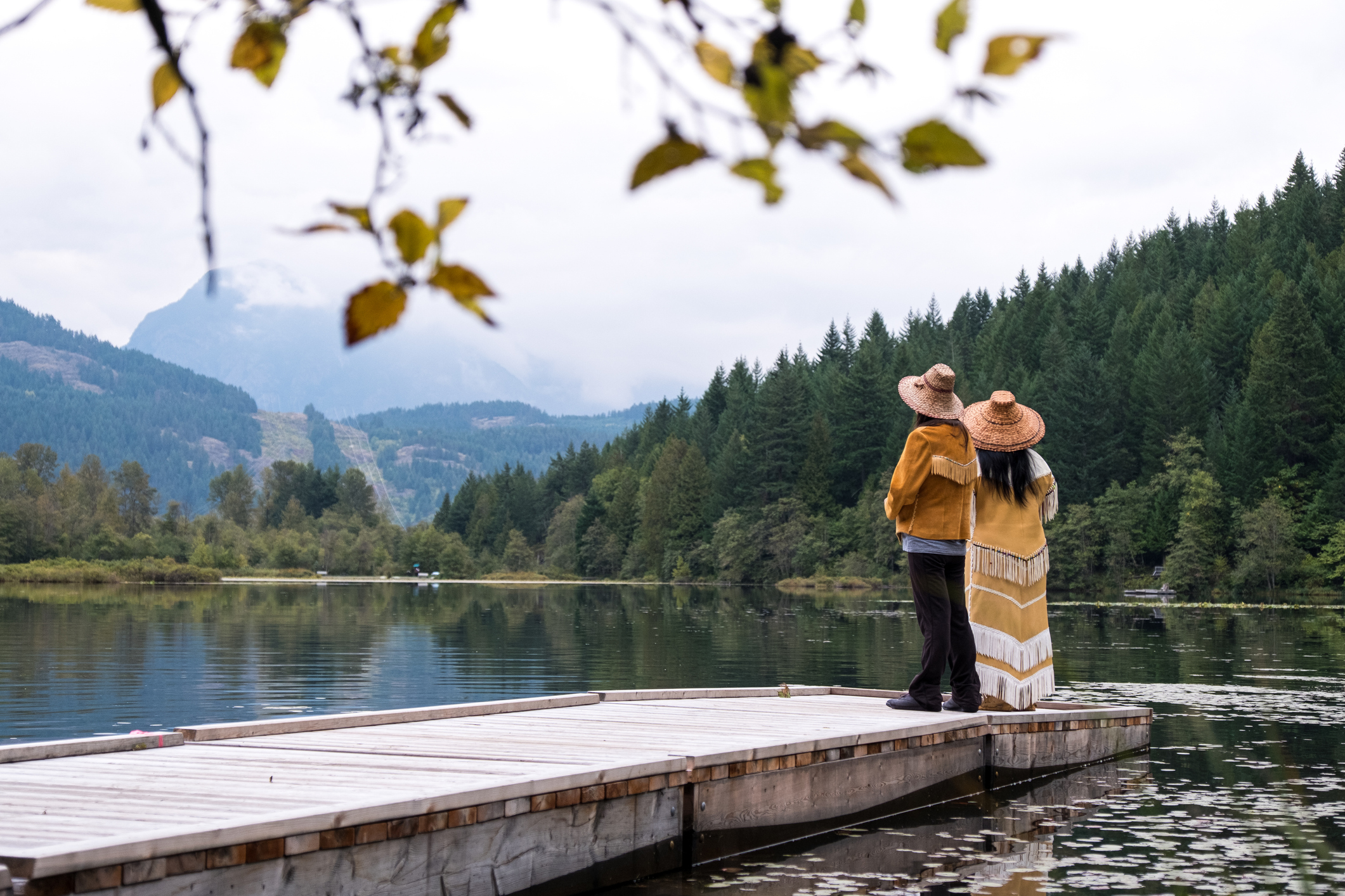 Two First Nations women dressed in traditional clothing on the shore of a lake. 