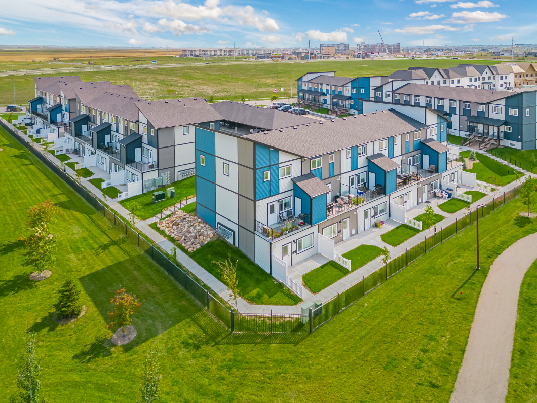 A wide-angle aerial view of the Aspen Heights development in Saskatoon. It consists of several two-storey townhouse complexes with blue and white exterior paneling and brown peaked roofs.