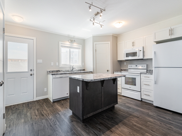 The interior of a home in the Aspen Heights development in Saskatoon. The image shows a brand new kitchen with a refrigerator, stove, island and counter space. It has dark wood floors and white cabinets and appliances.