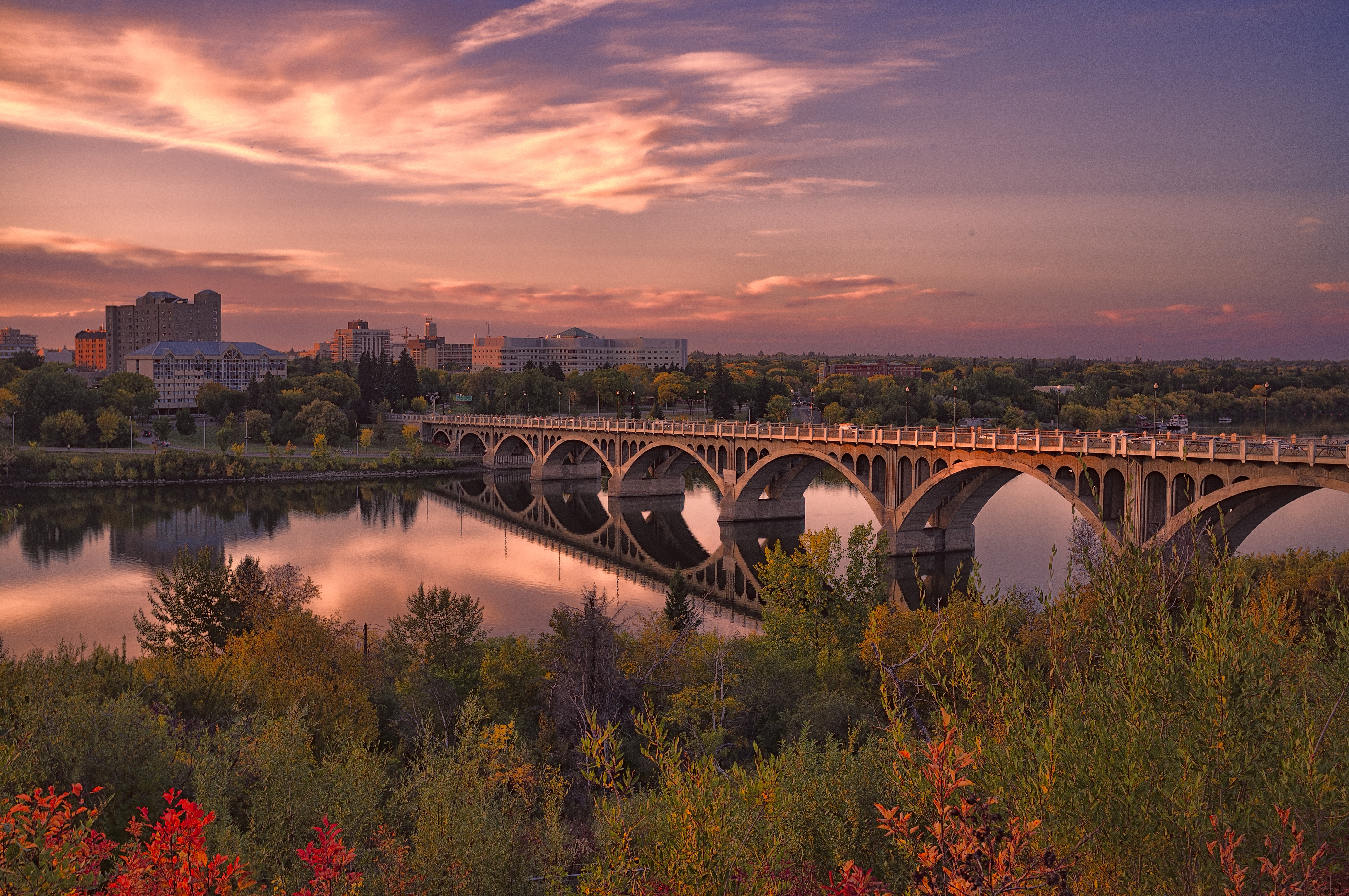 A calm river and bridge with greenery nearby and a cityscape beyond in the pink light of sunrise or sunset.