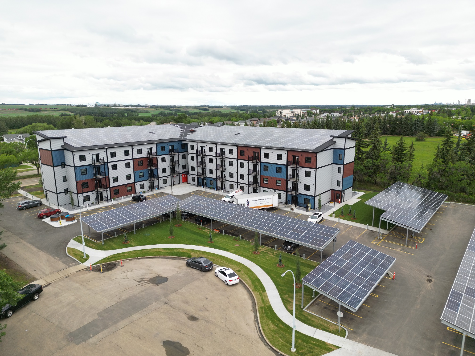 A wide-angle aerial view of the Heartland Housing Foundation's complex in Fort Saskatchewan, Alberta. It has two adjoined buildings, each four storeys, with an exterior designed with a blue, red and white mosaic pattern. The roofs are covered in solar panels, and the parking lot also features several large solar panels that each serve as a cover above 5-10 parking spots.