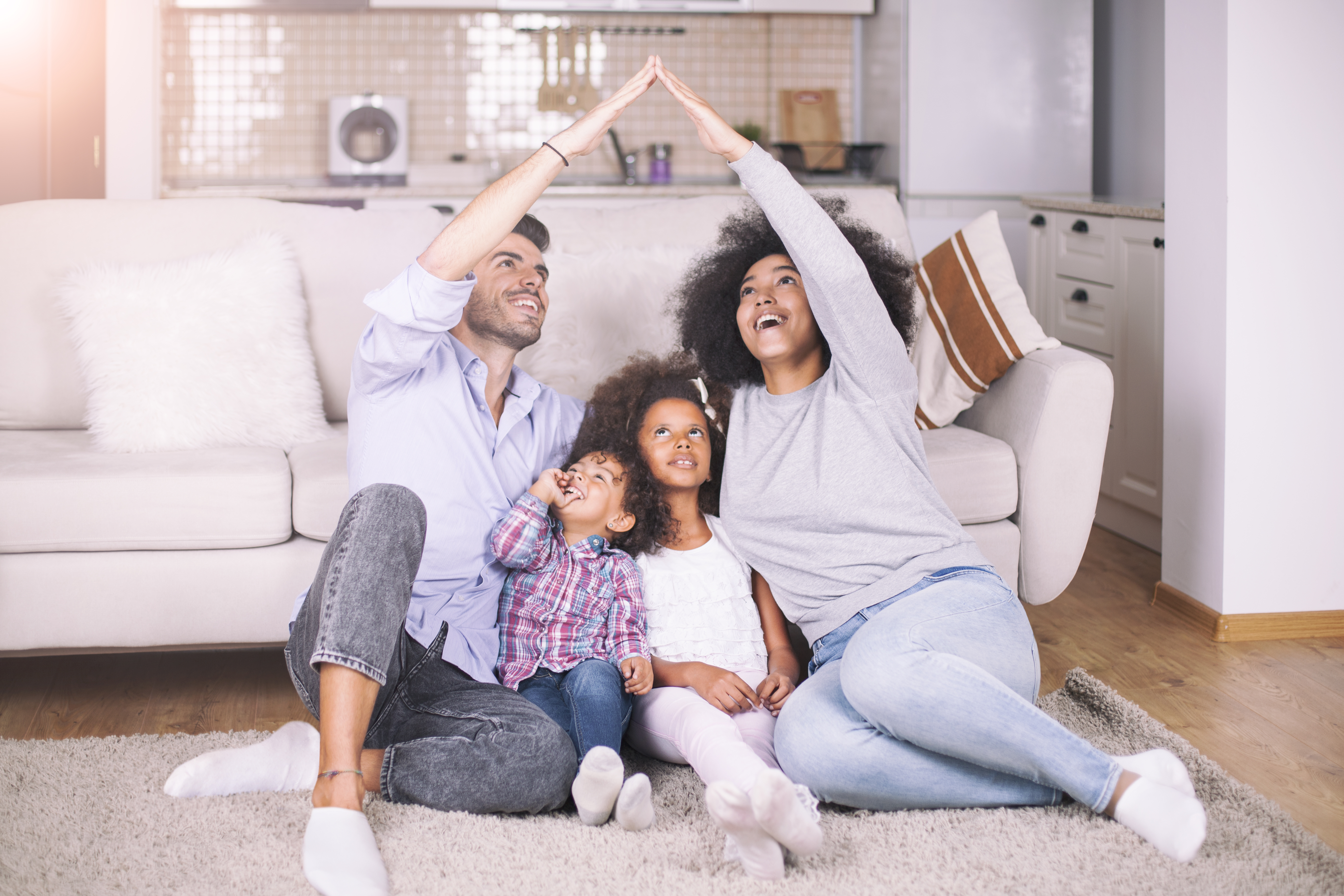 A family of four huddles together on the floor in front of their couch. The mother and father have their arms extended in the shape of the roof of a house over their two young children.