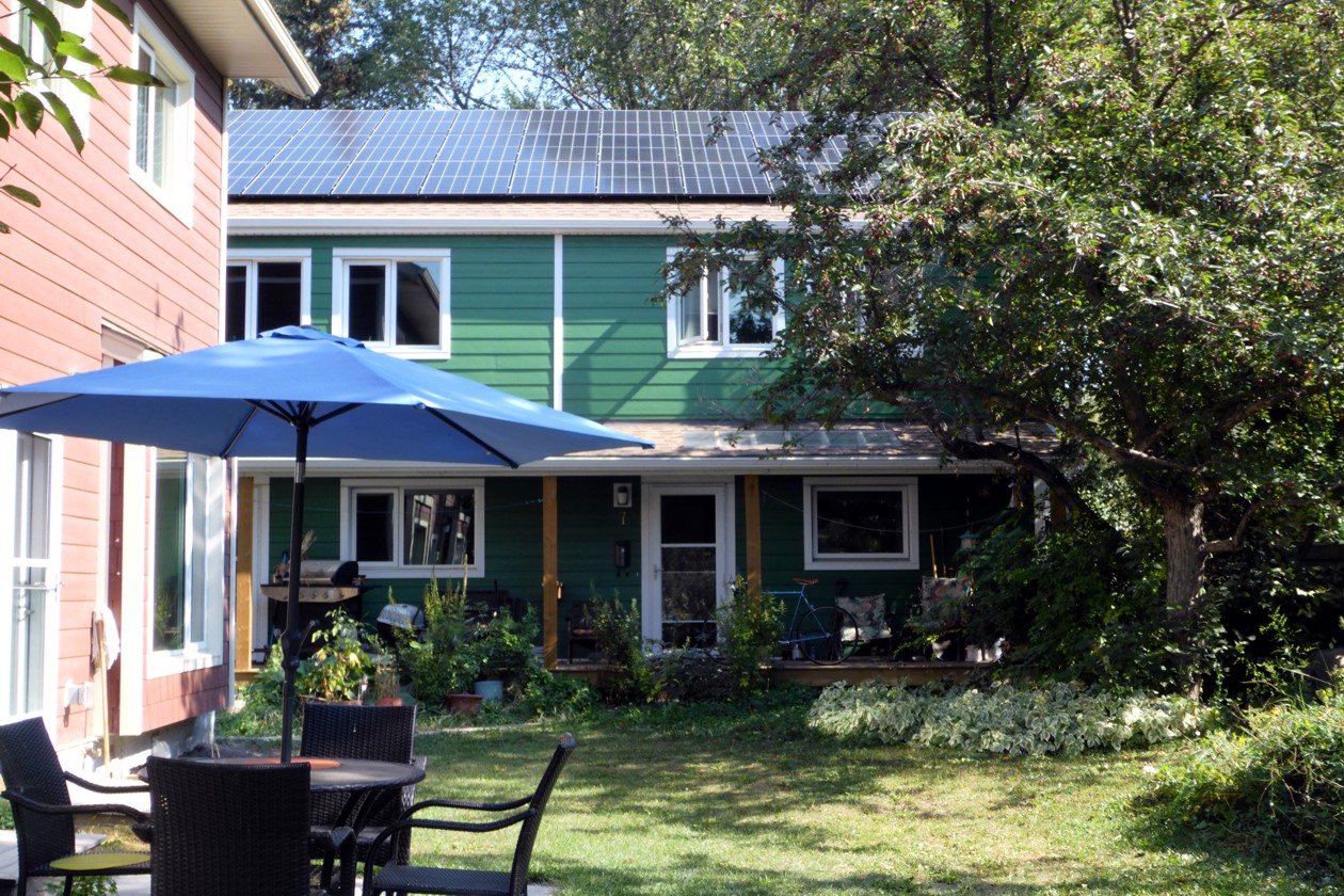 A view from the yard of a townhouse at Sundance Housing Co-operative in Edmonton. The two-storey home has a green exterior, windows and doors with white trim, and solar panels covering the entire roof. In the foreground, there is a grass lawn and a patio set with a blue umbrella.