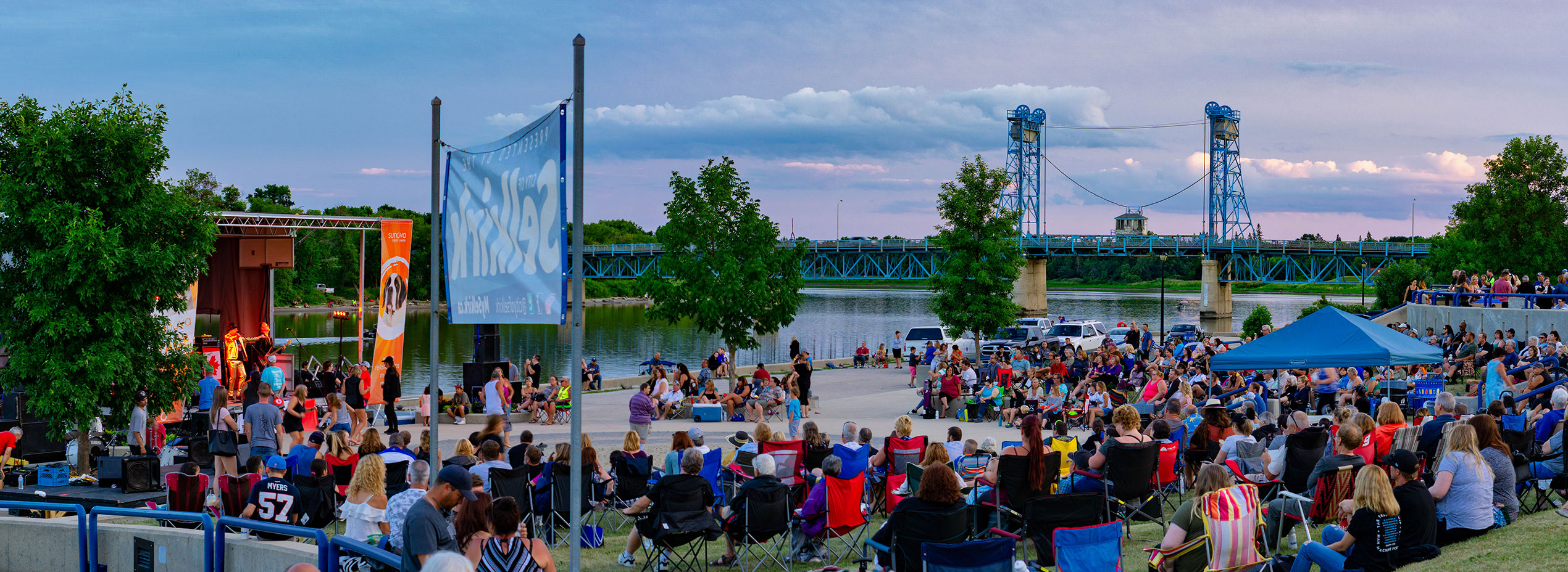 Photograph of a large group of people sitting on lawn chars around a stage, beside the Red River, with the Selkirk Bridge in the background and a large Selkirk sign in the foreground. 