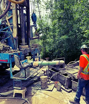 A worker in safety vest stands next to engineering equipment, with leafy green trees in background