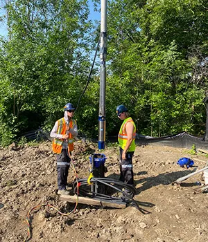Two workers in safety vests stand on either side of equipment, with leafy green trees in background