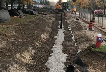 A long-angle view of a completed trench running alongside a road, backfilled with gravel and dirt. The area is marked by construction safety cones and surrounded by fences for security