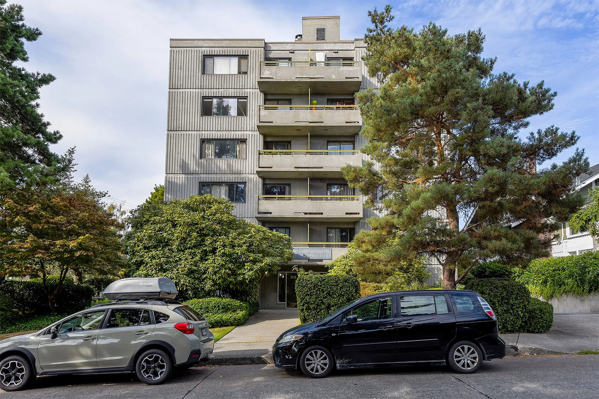 Side view of Brightside Community Home featuring a paved pathway leading to the entrance, two cars parked in front, and the building is nestled among hedges and trees