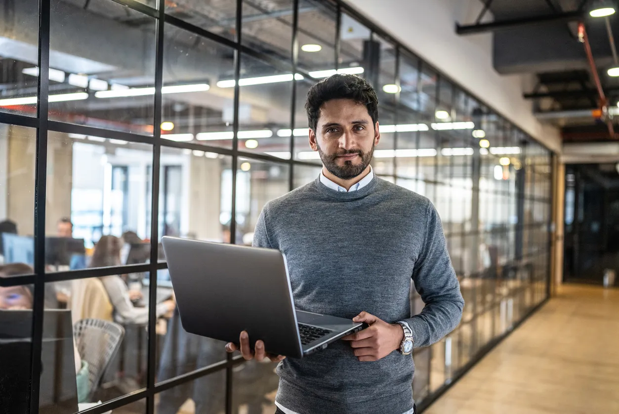 Man in sweater holding a laptop in a bright and sunny office.
