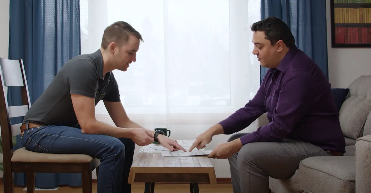 Two people in casual clothes sitting in a small home office, facing each other and looking over paperwork on a coffee table. 