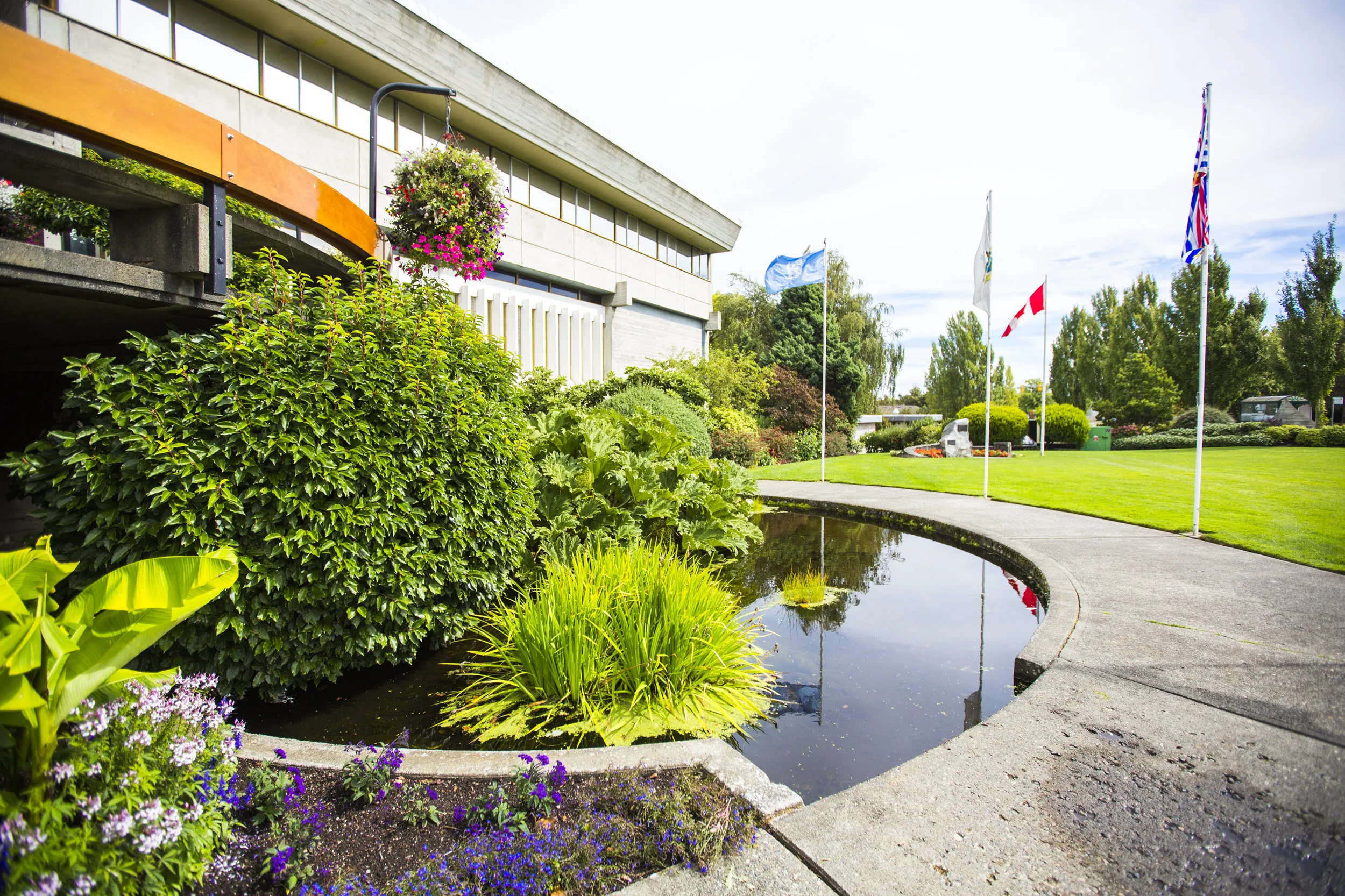 Exterior of a concrete building featuring a pond, grass, landscaping, flowers and flags as well as a circular pathway.