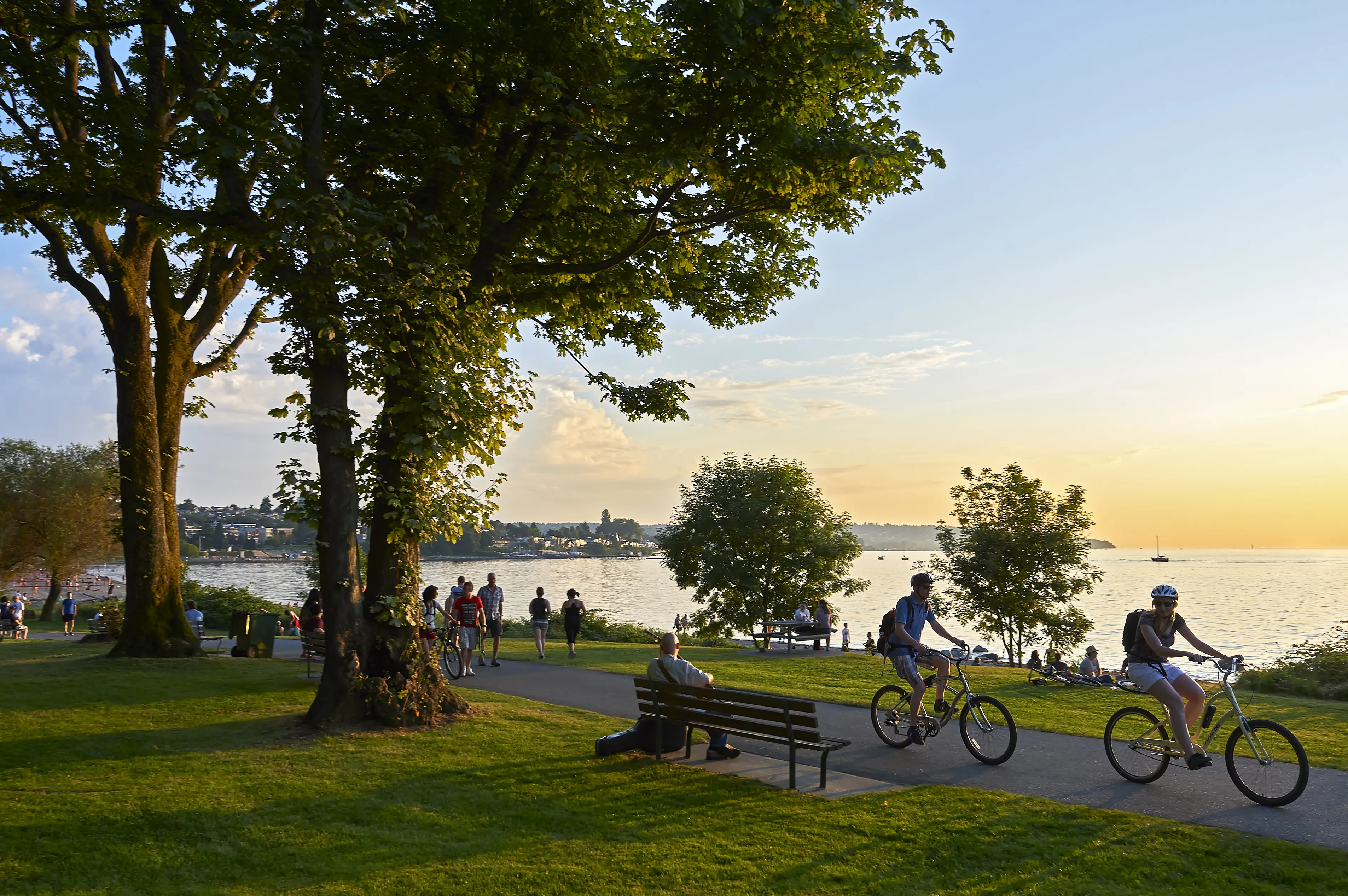 People biking, walking, and relaxing in a waterfront urban park at sunset. Some sit on benches or on the grass, while others enjoy the path along the shoreline.