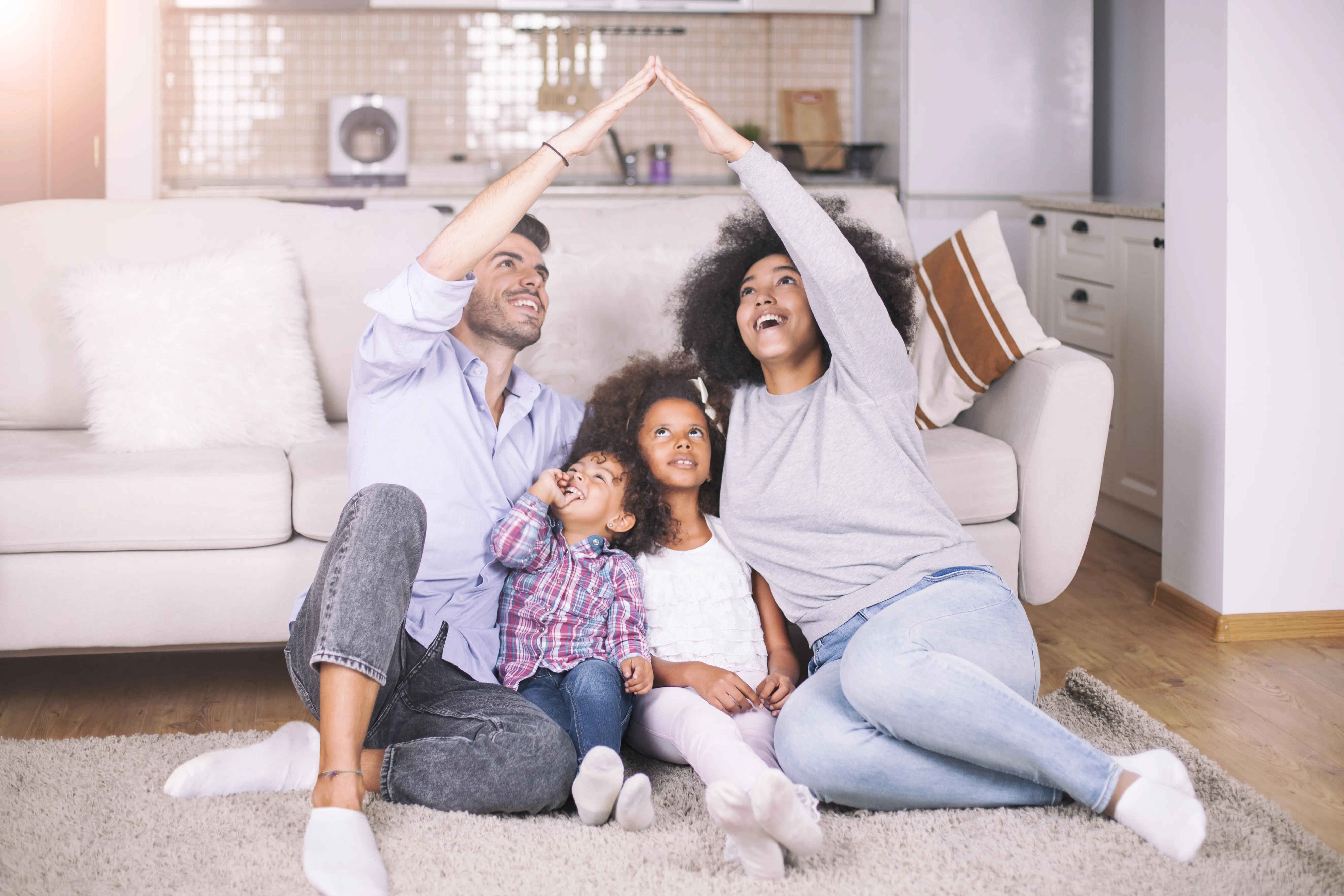 A family of four huddles together on the floor in front of their couch. The mother and father have their arms extended in the shape of the roof of a house over their two young children.