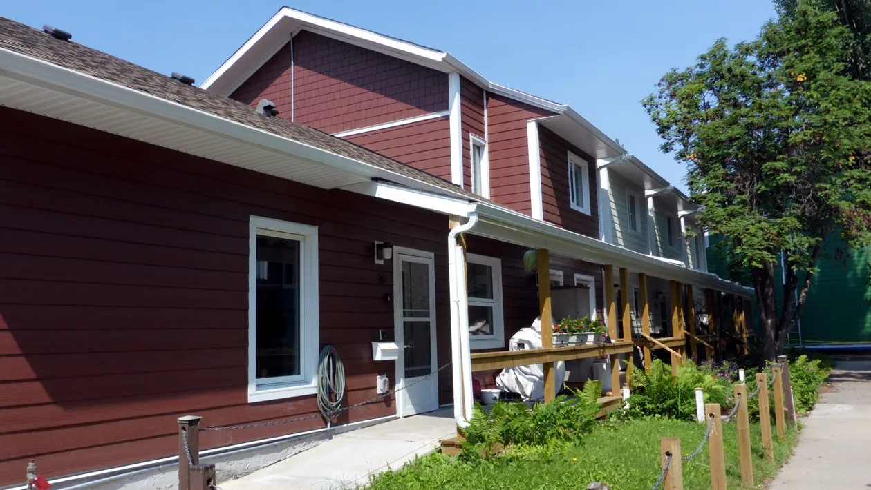 A ground-level view of a townhouse at Sundance Housing Co-operative in Edmonton. It has a dark red exterior with white trim, and there's greenery and a sidewalk out front.