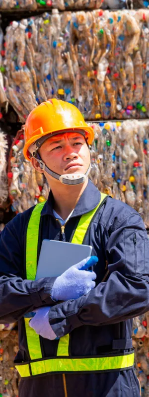 Man in yellow jump suit and yellow hard hat stands in front of a wall of colorful plastic waste pressed into large bricks.