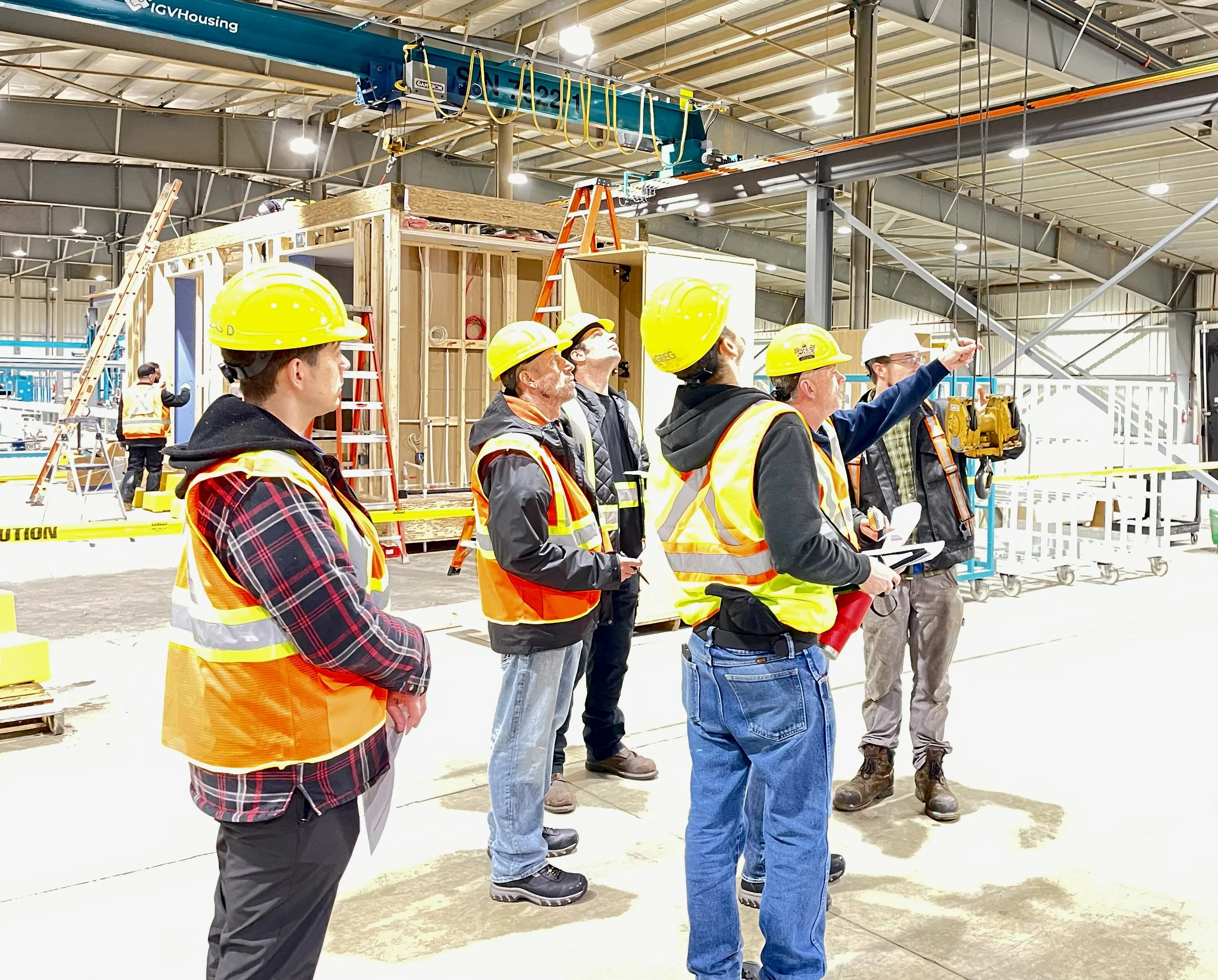 Group of workers in a brightly lit industrial setting wearing yellow hard hard and orange safety vests.