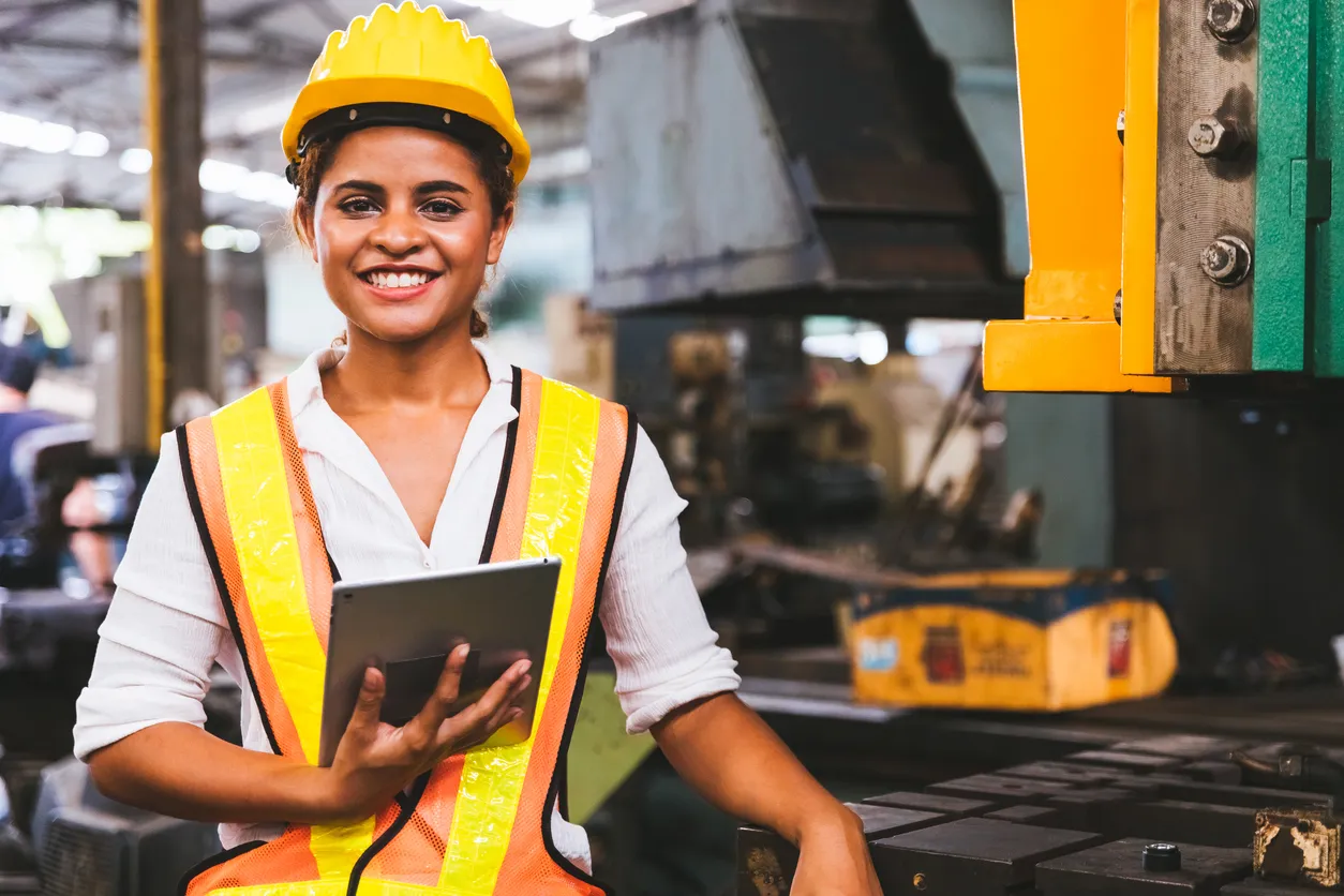 Woman in hard hat and safety vest smiles in front of industrial machinery. 