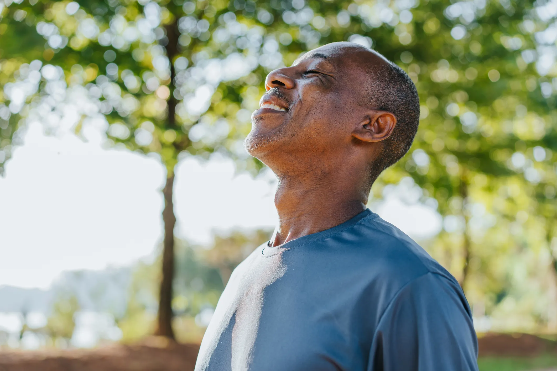 A person outside on a sunny day, with their head tilted back, eyes closed, and a smile on their face, savoring the air purified by the trees.